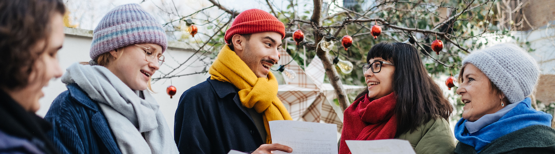 Fünf Nachbar:innen in Winterkleidung stehen draußen vor einem weihnachtlich geschmückten Baum mit Zetteln in der Hand und singen Weihnachtslieder