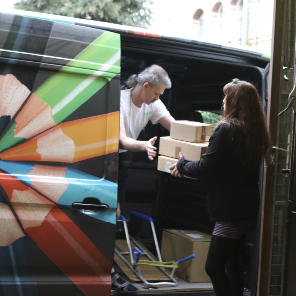 2 Mitarbeitende vom M&W Bürobedarf laden Kartons in den mit Buntstiften verzierten Lieferwagen.