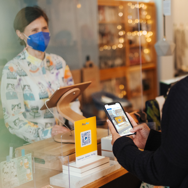 Eine Kundin scannt den PayPal QR-Code an der Kasse mit dem Handy. Ladenbesitzerin Sabine steht hinter einer transparenten Wand und freut sich, dass sie kontaktlos und einfach bezahlt wird.