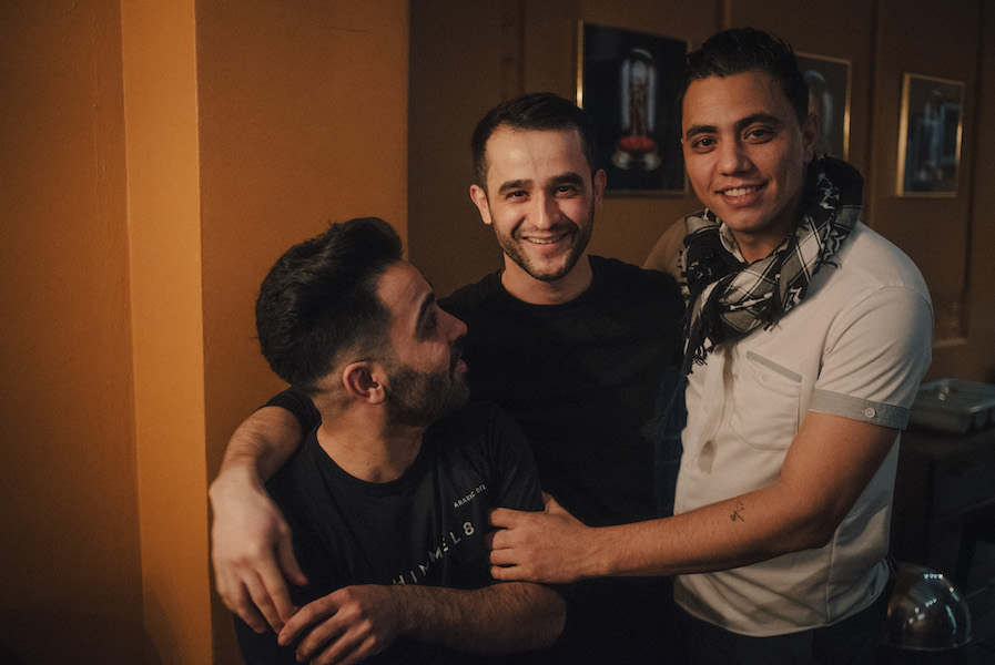 Drei junge Männer umarmen sich, zwei lächeln in die Kamera