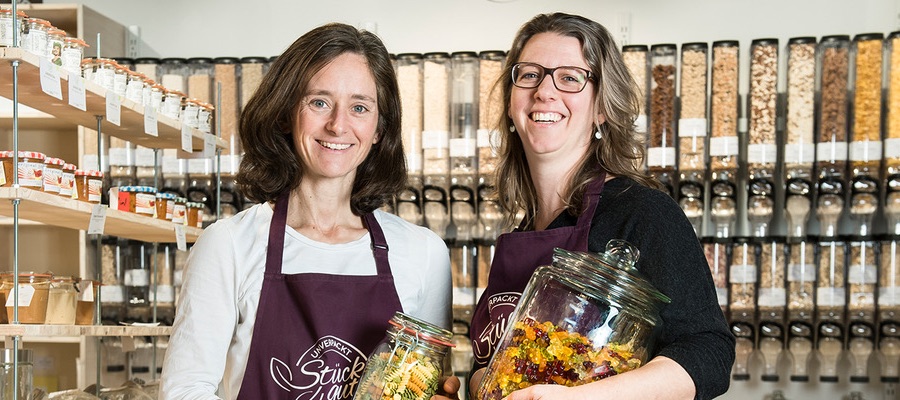 Sonja und Insa stehen lächeln in ihrem Laden, vor einem Unverpacktregal. In den Händen halten sie zwei Gläser mit unverpackten Gummibärchen und Nudeln und tragen eine Schürze mit dem Stückgut Logo