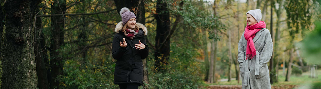 Zwei Frauen in Winterkleidung spazieren auf Abstand zusammen durch den Wald und unterhalten sich