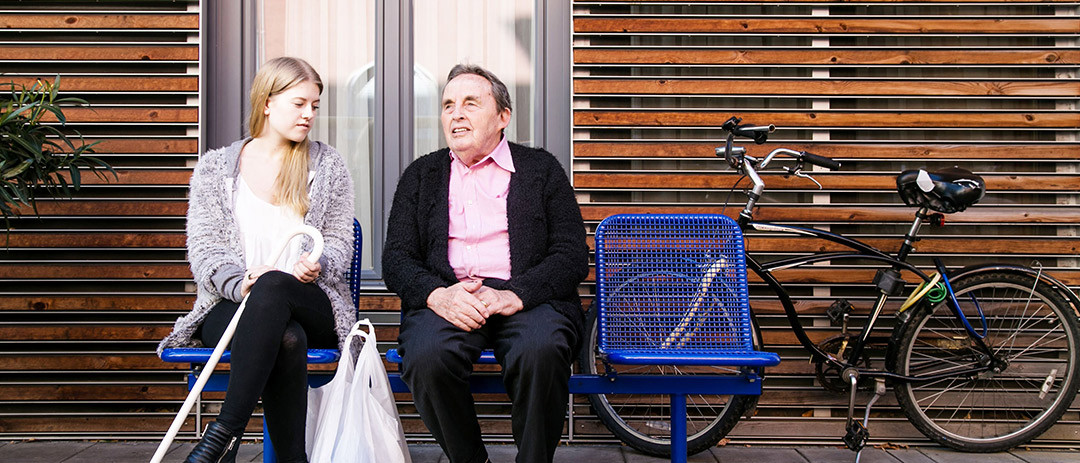 Junge Frau und älterer Mann sitzen gemeinsam auf einer Bank und unterhalten sich.