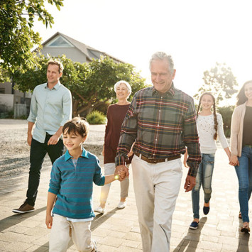 Familie spaziert durch sonnige Nachbarschaft, Großvater hält Enkel im Vordergrund an der Hand.