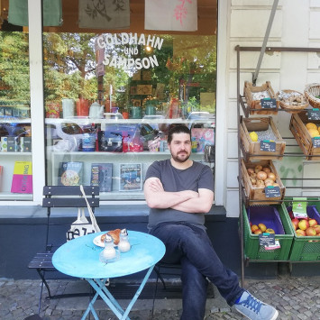 Nebenan.de Nachbar Philipp sitzt auf Stuhl vor seinem Lieblingsladen neben Obst- und Gemüsekisten