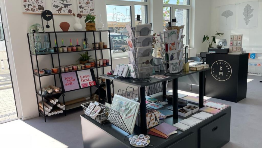 Ein Blick in den im modernen weiß und schwarz gehaltenen Laden zeigt ein buntes Sortiment aus Postern, Keramik, Notizblöcken, Postkarten und vielem mehr. Auf der Ladentheke steht das runde Logo des Kollektiv Mainz