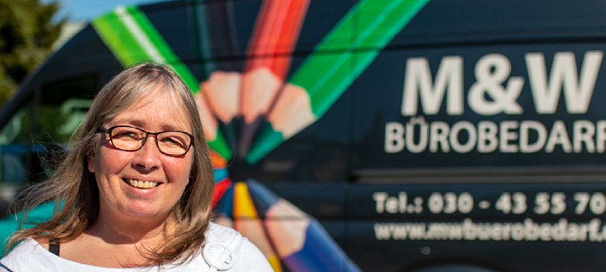 Birgit steht mit strahlendem Lächeln vor einem ihrer Transporter. Dieser trägt die Aufschrift M&W Bürobedarf und ist mit Buntstiften verziert. Birgit hat blondes Haar trägt eine Brille und eine weiße Bluse.