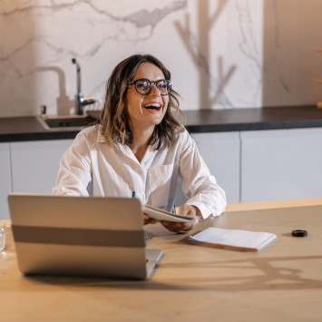 Frau mit Brille sitzt lachend vor Laptop.