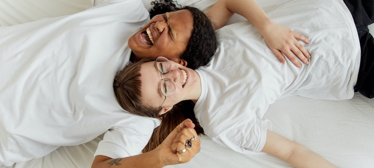 Zwei junge Frauen liegen in weißen T-Shirts auf einem weißen Bett und lachen herzhaft.