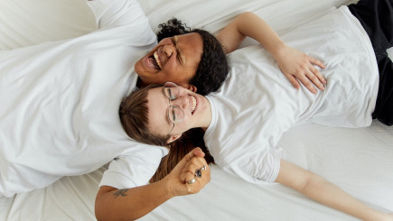 Zwei junge Frauen liegen in weißen T-Shirts auf einem weißen Bett und lachen herzhaft.