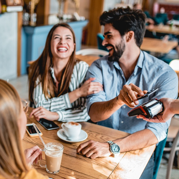 Freundesgruppe sitzt lachend um Café; ein junger Mann hält sein Smartphone über das Zahlungsgerät.