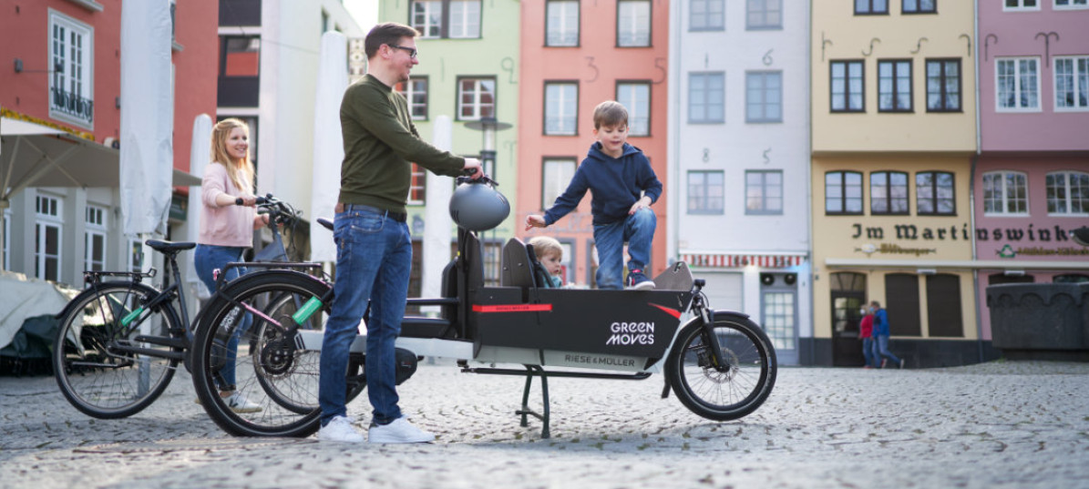 Mit dem E-Lastenrad transportiert ein Vater seine Kinder ber den gepflasterten Marktplatz. Seine Frau hat ein E-Bike gewählt