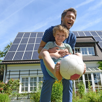 Vor einem Haus mit Solarpanelen auf dem Dach hebt ein Mann lachend ein Kind hoch, der Ball zwischen den Füßen hält.