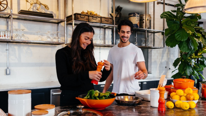 Junge Frau und Mann stehen lachend an einer Kücheninsel und bereiten Essen vor.