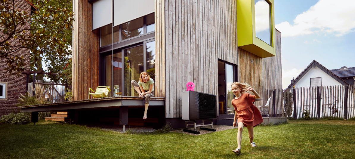 Kinder spielen im Garten eines modernen Holzhauses, im Hintergrund neben dem Haus steht die Daikin Wärmepumpe auf der ein kleiner pinker Octopus sitzt