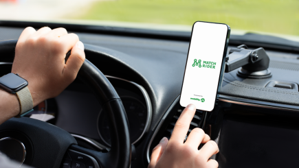 Neben dem Fahrersitz eines PKW ist ein Smartphone angebracht. Mit der rechten Hand öffnet eine fahrende Person die Match Rider App, deren grün weißes Logo auf dem Bildschirm erscheint.