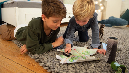 Zwei Jungs liegen auf einem Teppich und schauen mit einer Lupe auf eine selbst gezeichnete Landkarte.