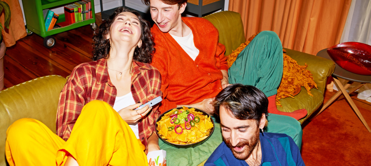 Lachend sitzen drei junge Personen vor und auf dem Sofa und genießen Nachos mit SimplyV veganem Käse