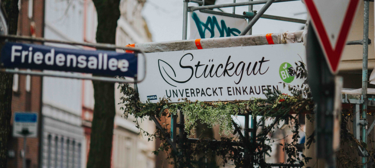 Neben dem Straßenschild der Friedensallee in Hamburg hängt das Ladenschild von Stückgut. In geschwungenen Buchstaben steht dort 