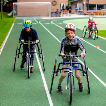 Dreit Kinder mit Helm nutzen ihre Larfräder auf einer Rennstrecke, die ihnen das Rennen trotz Gehbehinderung ermöglichen.