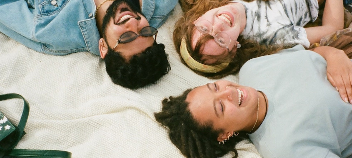 Drei Menschen liegen lachend auf einer Decke im Gras, die Köpfe zueinander.