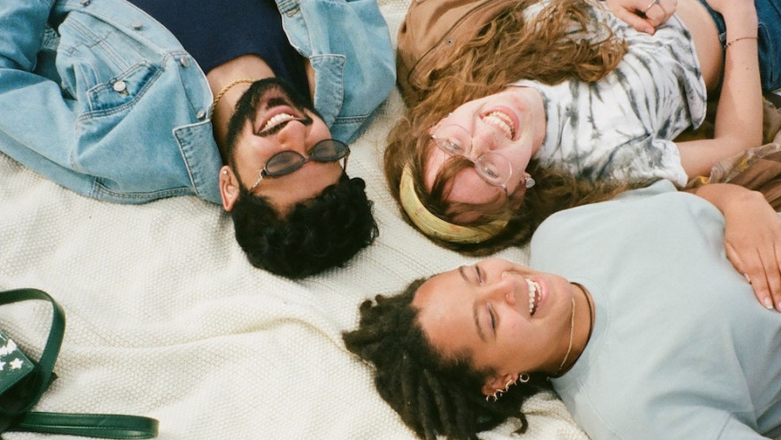 Drei Menschen liegen lachend auf einer Decke im Gras, die Köpfe zueinander.