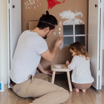 Vater und Tochter bemalen gemeinsam ein Pappkartonhaus.