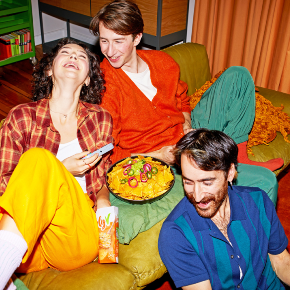 Lachend sitzen drei junge Personen vor und auf dem Sofa und genießen Nachos mit SimplyV veganem Käse